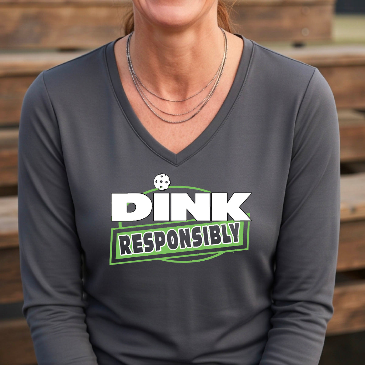 A woman wearing a gray long-sleeve V-neck t-shirt with a pickleball graphic that reads 'DINK RESPONSIBLY' in white and green,.