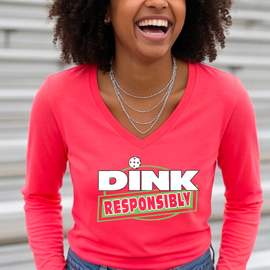 A woman wearing a bright coral long-sleeve V-neck t-shirt with a pickleball graphic that reads 'DINK RESPONSIBLY' in white and green,.