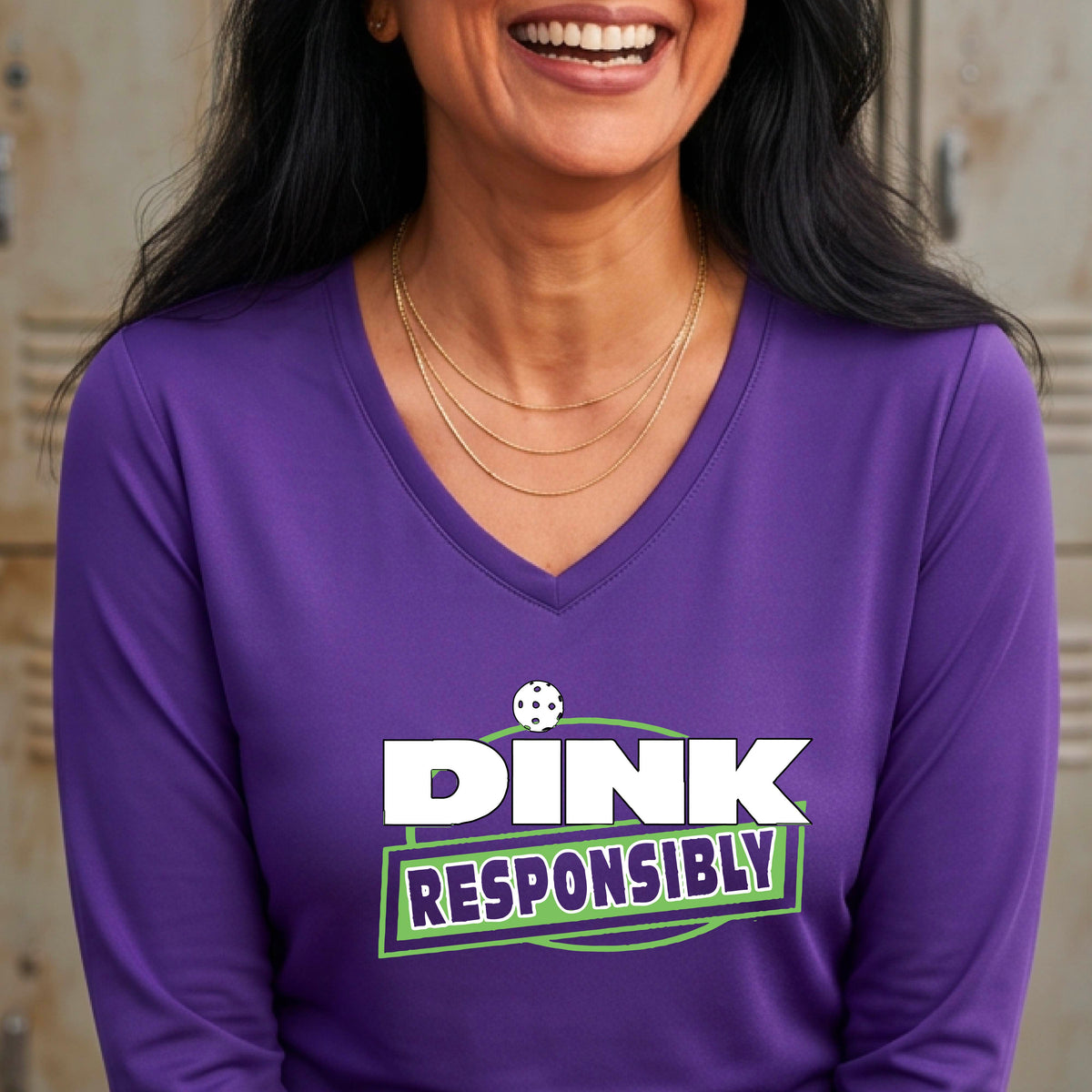 A woman wearing a purple long-sleeve V-neck t-shirt with a pickleball graphic that reads 'DINK RESPONSIBLY' in white and green,.