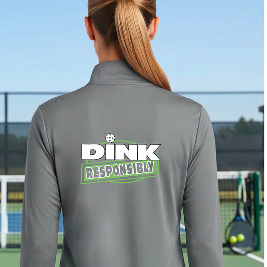 A woman viewed from behind wearing a gray athletic quarter-zip jacket with a 'DINK RESPONSIBLY' logo printed on the back, set against an outdoor pickleball court.