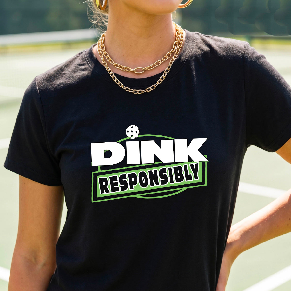 A woman wearing a pickleball black short-sleeve graphic t-shirt with the text 'DINK RESPONSIBLY' in white and green.