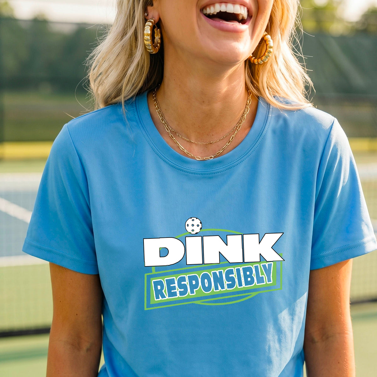 A woman wearing a pickleball light blue short-sleeve graphic t-shirt with the text 'DINK RESPONSIBLY' in white and green.