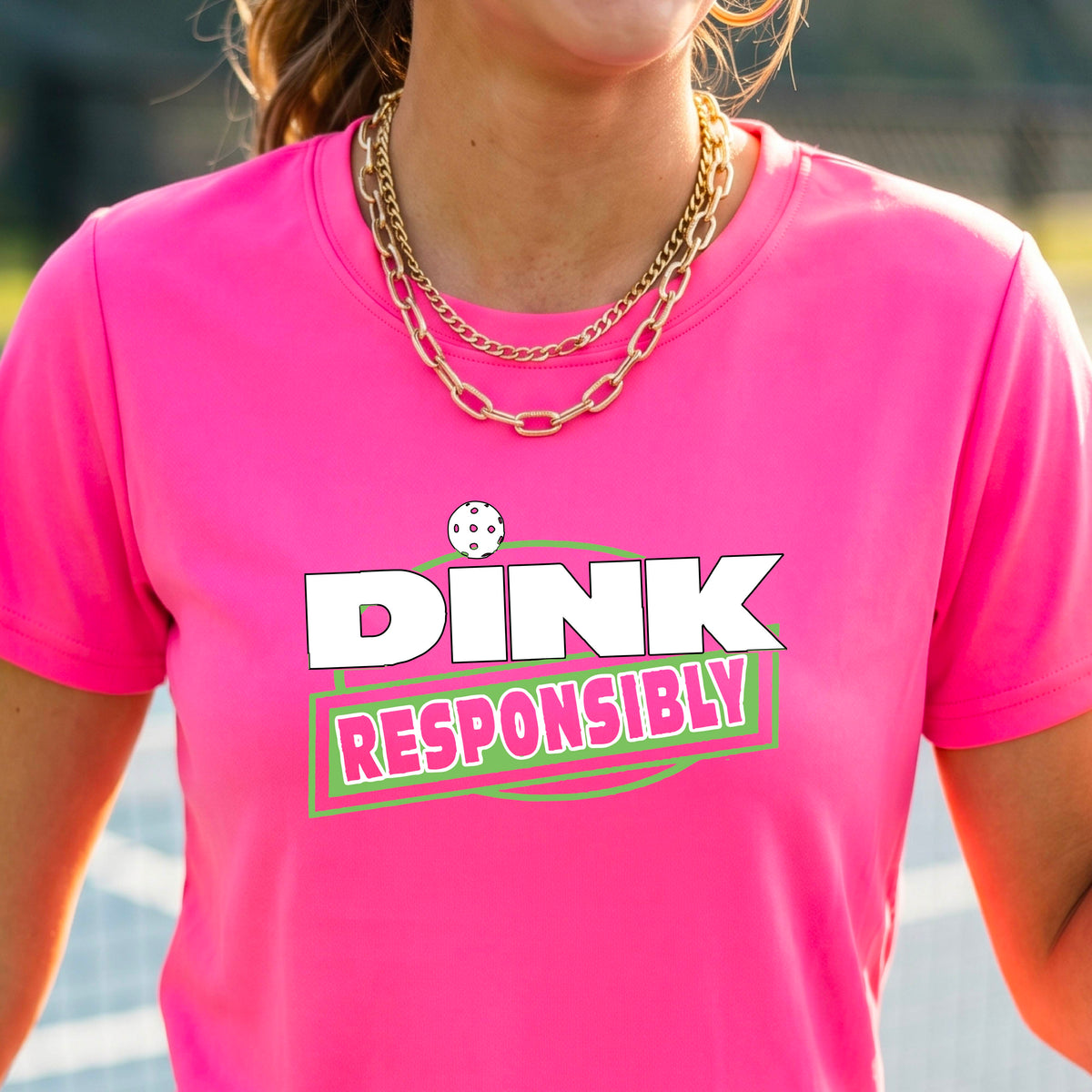 A woman wearing a pickleball pink short-sleeve graphic t-shirt with the text 'DINK RESPONSIBLY' in white and green.