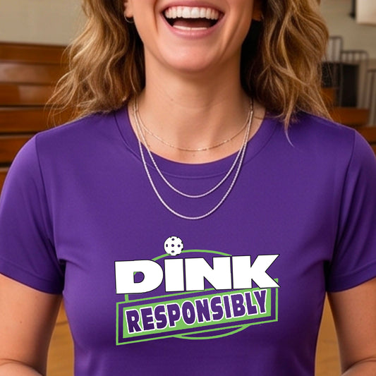 A woman wearing a pickleball purple short-sleeve graphic t-shirt with the text 'DINK RESPONSIBLY' in white and green.