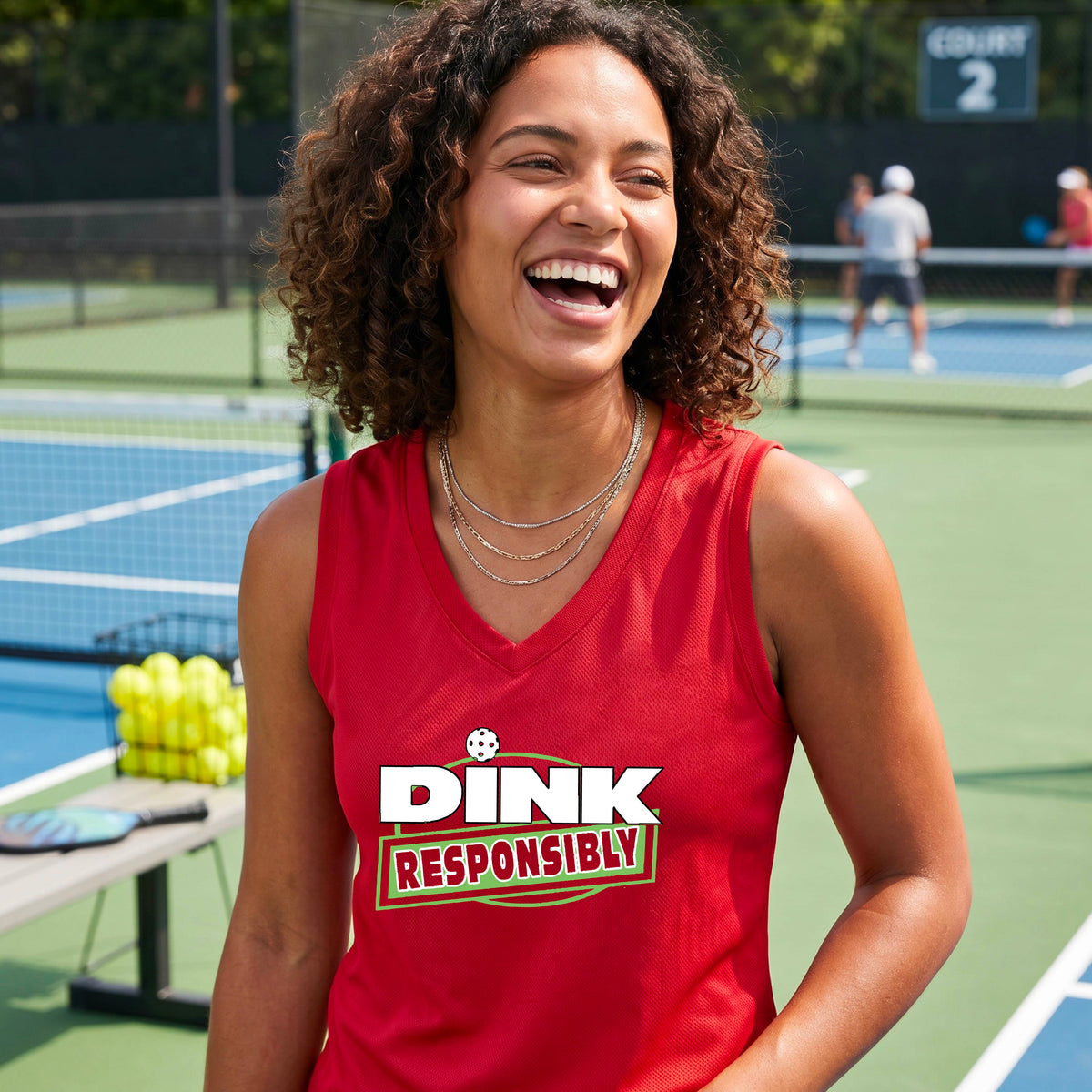 A smiling woman on a pickleball court wearing a red sleeveless V-neck athletic tank top with a green and white 'DINK Responsibly' graphic on the front,.