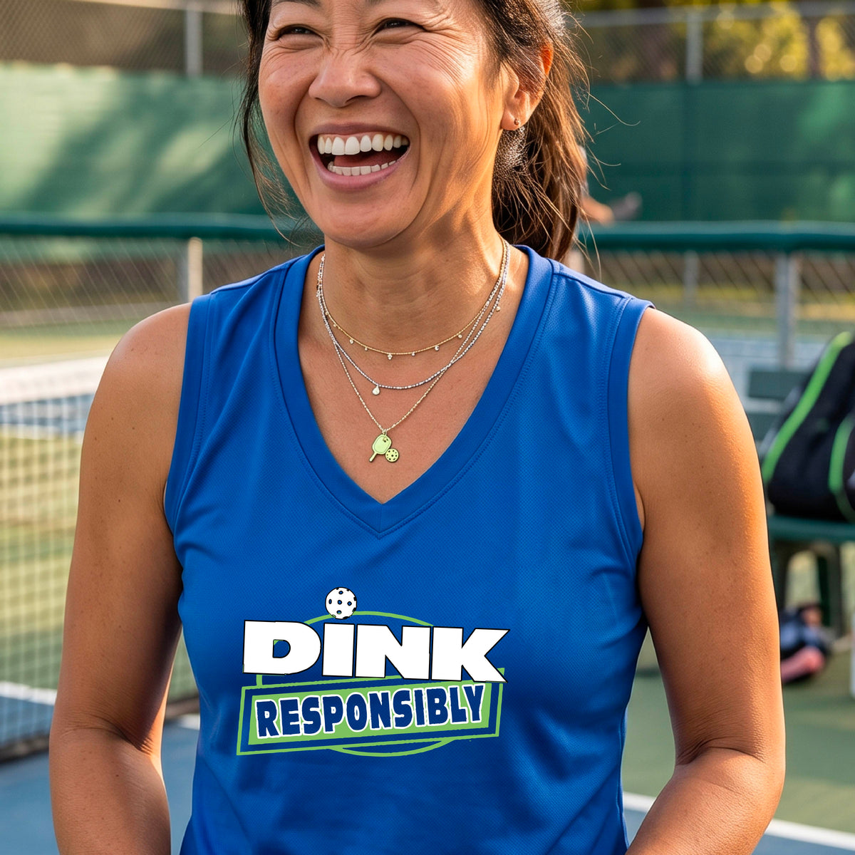 A smiling woman on a pickleball court wearing a royal blue sleeveless V-neck athletic tank top with a green and white 'DINK Responsibly' graphic on the front,.