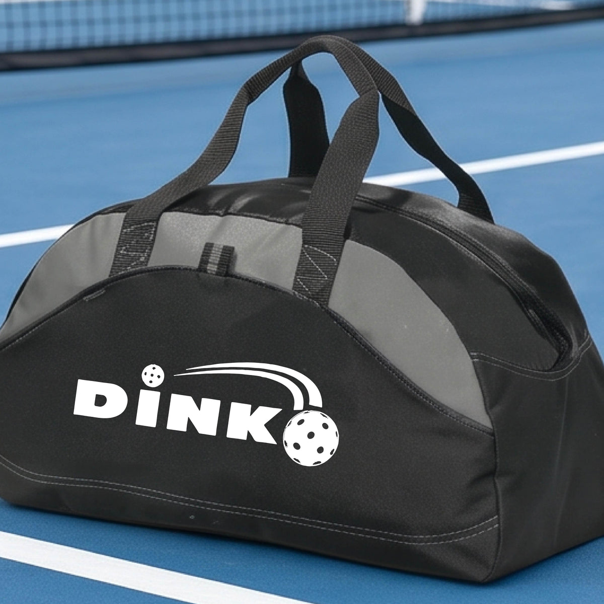 A close up of a black and gray pickleball  duffel bag with a pickleball design of text 'DINK' with a white pickleall in flight, on a pickleball court .