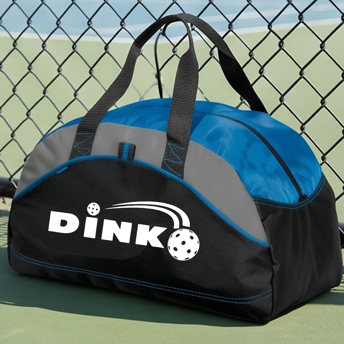 A close up of a royal blue, black and gray pickleball  duffel bag with a pickleball design of text 'DINK' with a white pickleall in flight, on a pickleball court .