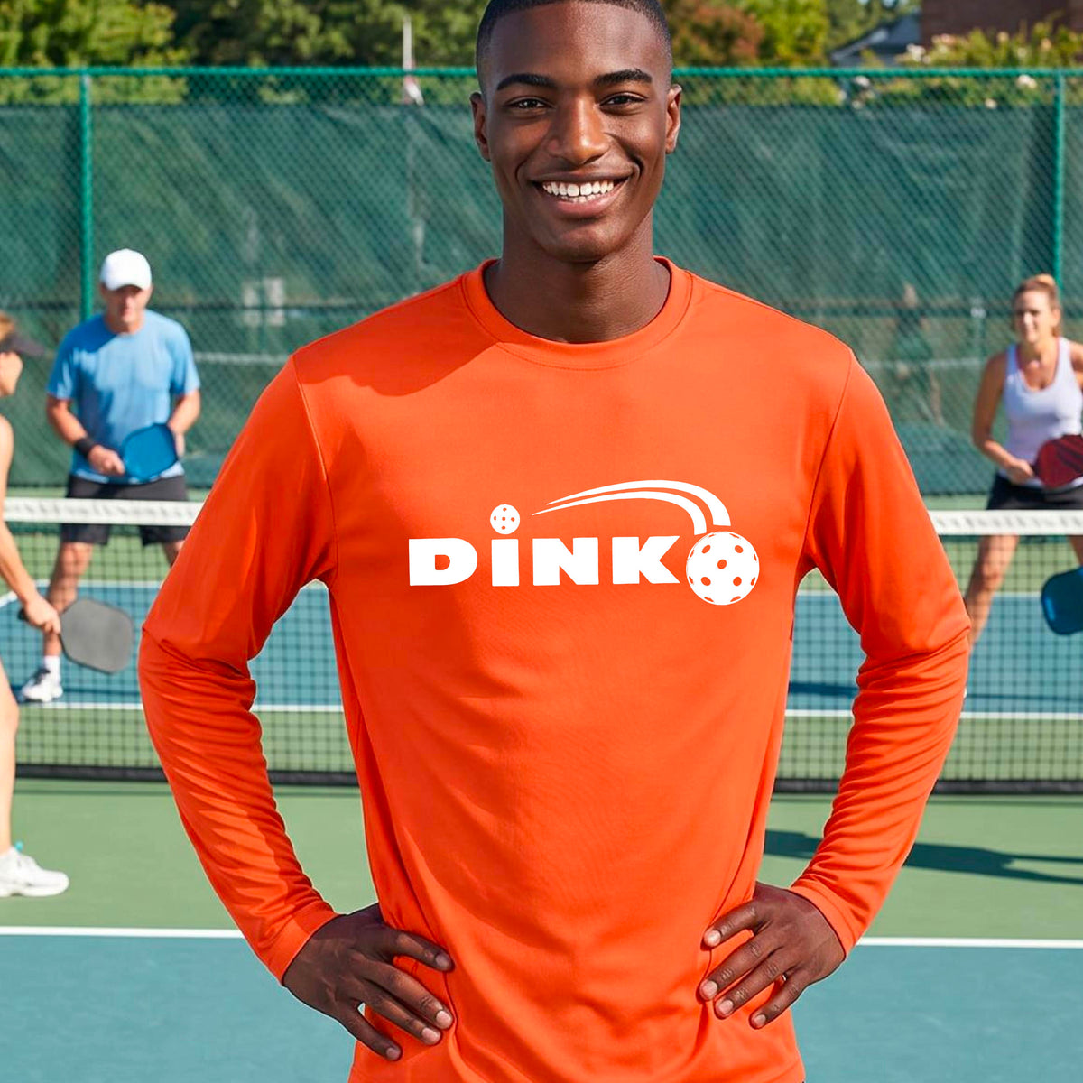 A man wearing a bright orange long-sleeve t-shirt with a pickleball design of text 'DINK' with a white pickleall in flight, standing on a pickleball court.
