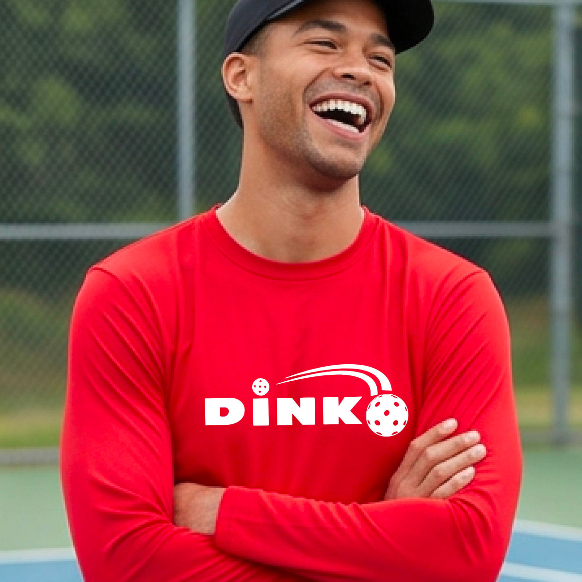 A man wearing a red long-sleeve t-shirt with a pickleball design of text 'DINK' with a white pickleall in flight, standing on a pickleball court.