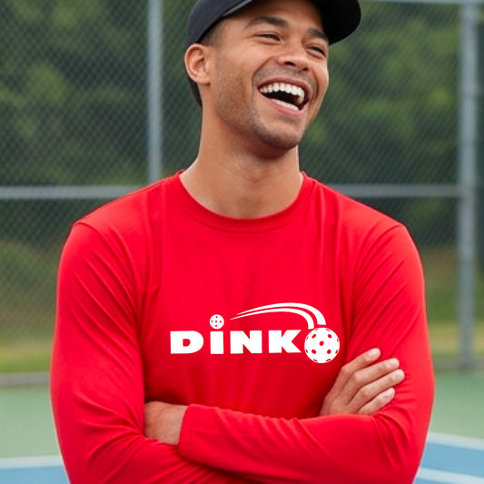 A man wearing a red long-sleeve t-shirt with a pickleball design of text 'DINK' with a white pickleall in flight, standing on a pickleball court.