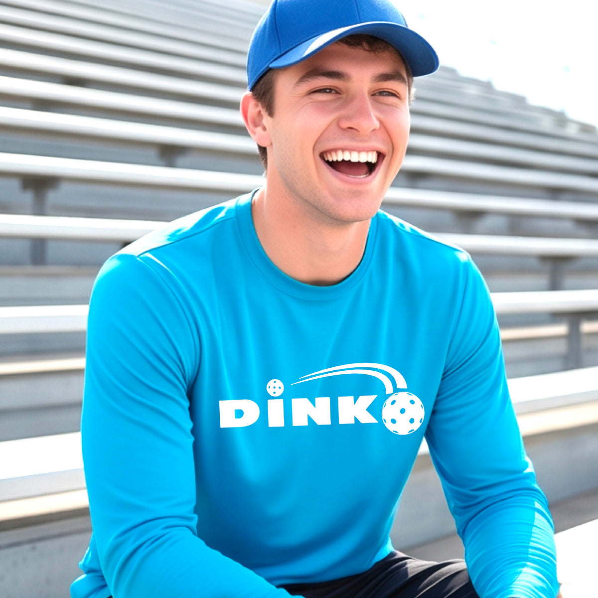 A man wearing a bright blue long-sleeve t-shirt with a pickleball design of text 'DINK' with a white pickleall in flight, standing on a pickleball court.