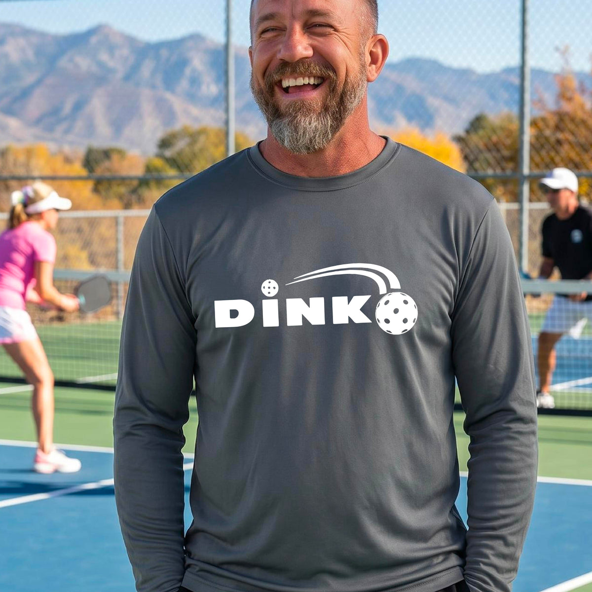 A man wearing a gray long-sleeve t-shirt with a pickleball design of text 'DINK' with a white pickleall in flight, standing on a pickleball court.