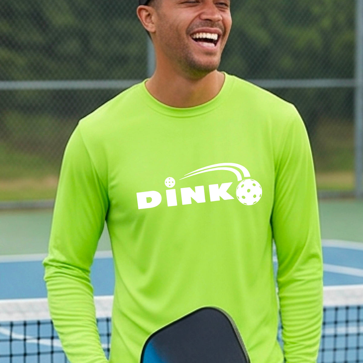 A man wearing a lime green long-sleeve t-shirt with a pickleball design of text 'DINK' with a white pickleall in flight, standing on a pickleball court.