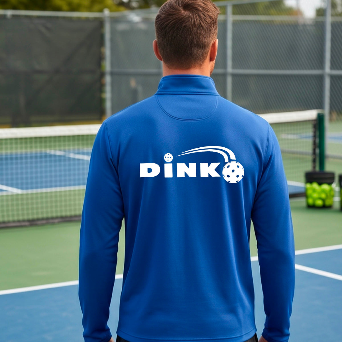A man viewed from behind wearing a royal  blue long-sleeve quarter-zip pullover with a white DINK logo and a pickleball in flight printed across the back, standing on an outdoor pickleball  court.