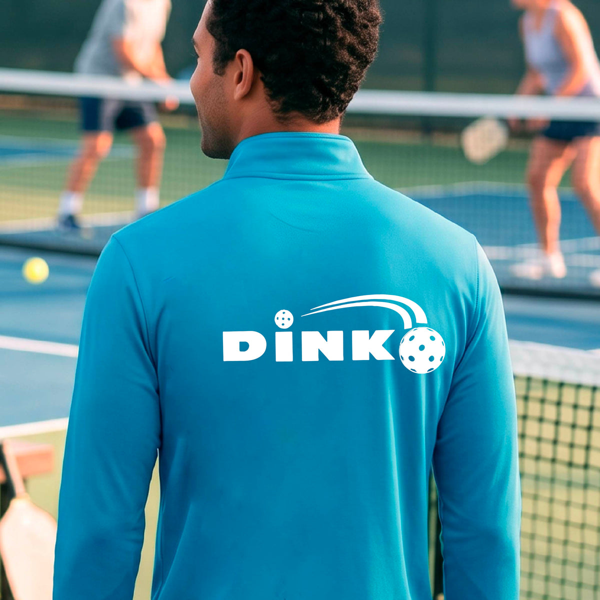 A man viewed from behind wearing a bright blue long-sleeve quarter-zip pullover with a white DINK logo and a pickleball in flight printed across the back, standing on an outdoor pickleball  court.