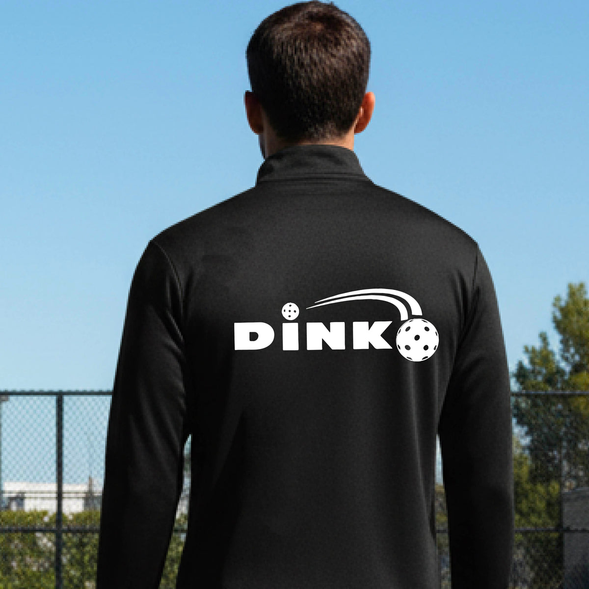 A man viewed from behind wearing a black long-sleeve quarter-zip pullover with a white DINK logo and a pickleball in flight printed across the back, standing on an outdoor pickleball  court.