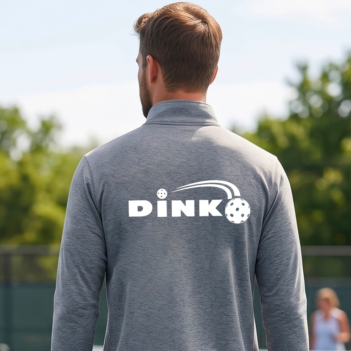 A man viewed from behind wearing a gray long-sleeve quarter-zip pullover with a white DINK logo and a pickleball in flight printed across the back, standing on an outdoor pickleball  court.