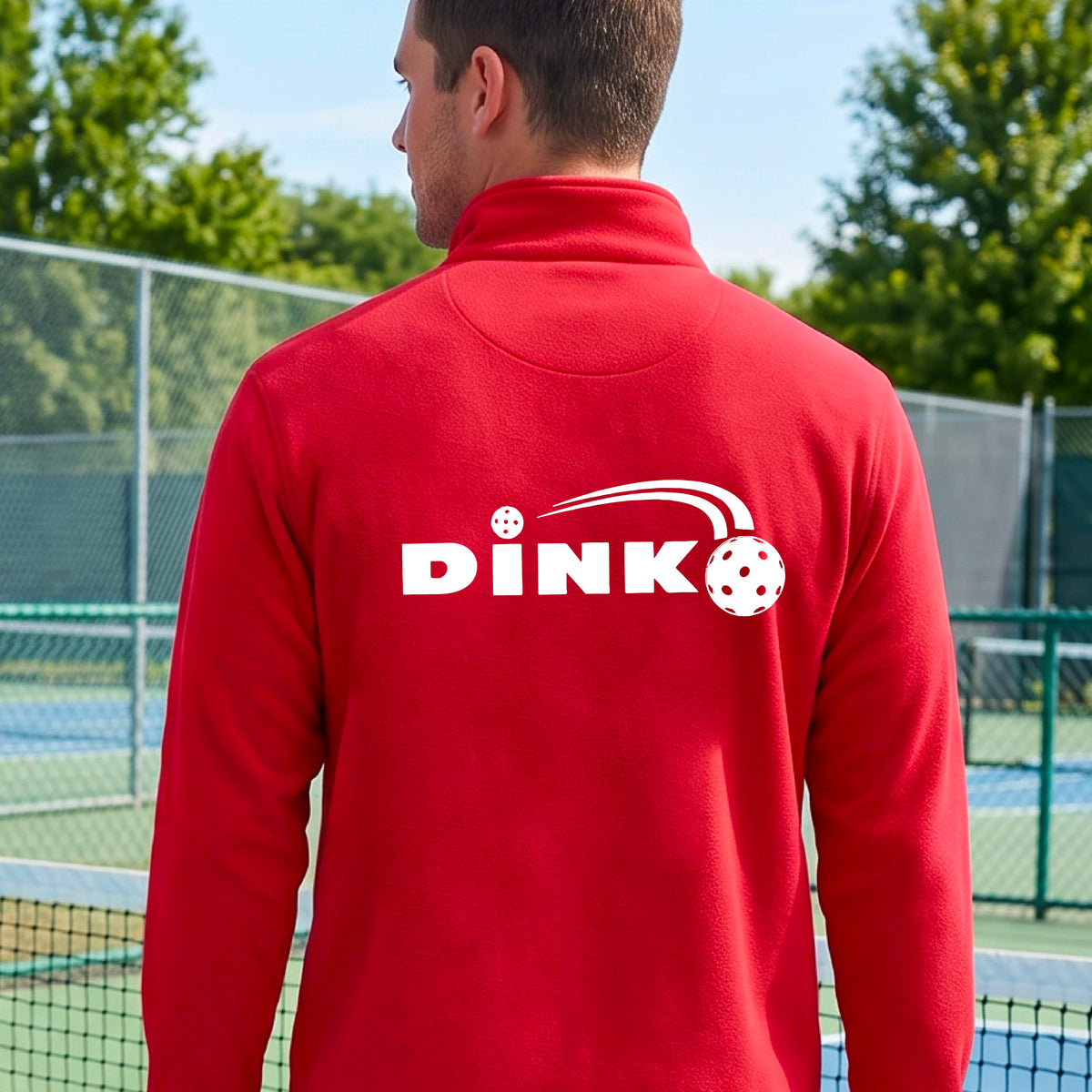 A man viewed from behind wearing a bright red long-sleeve quarter-zip pullover with a white DINK logo and a pickleball in flight printed across the back, standing on an outdoor pickleball  court.