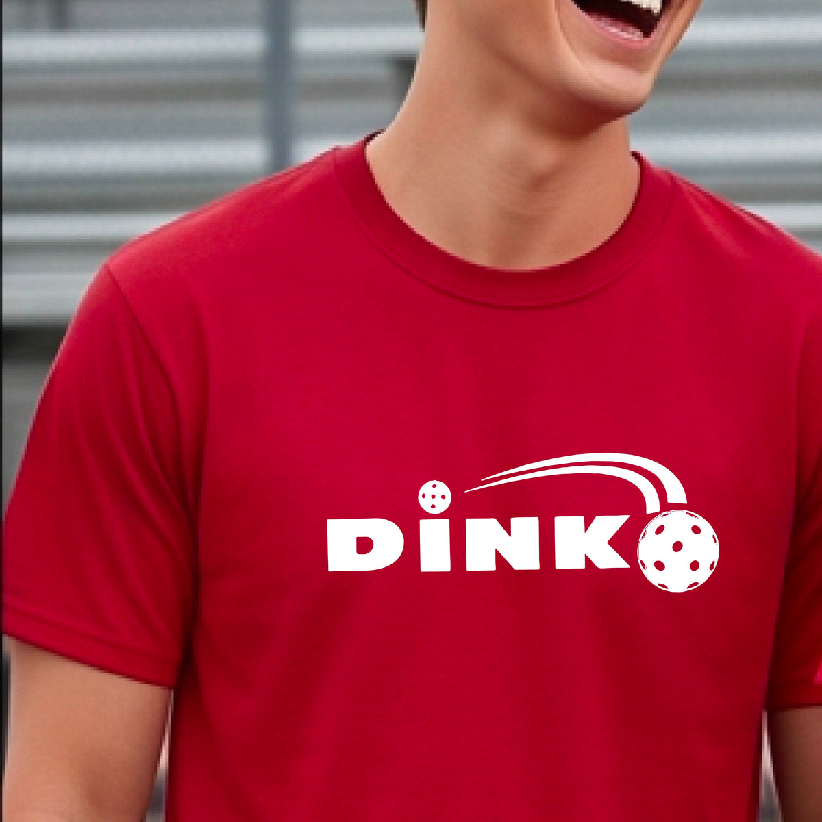 A man wearing a red short-sleeve t-shirt with a pickleball design of text 'DINK' with a white pickleall in flight, standing on a pickleball court.