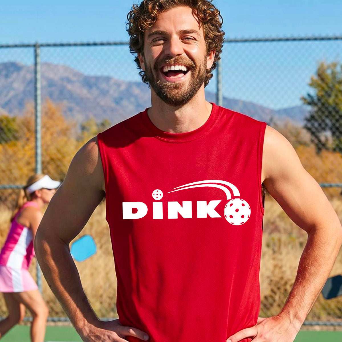 A man wearing a red sleeveless t-shirt with a pickleball design of text 'DINK' with a white pickleall in flight, standing on a pickleball court.