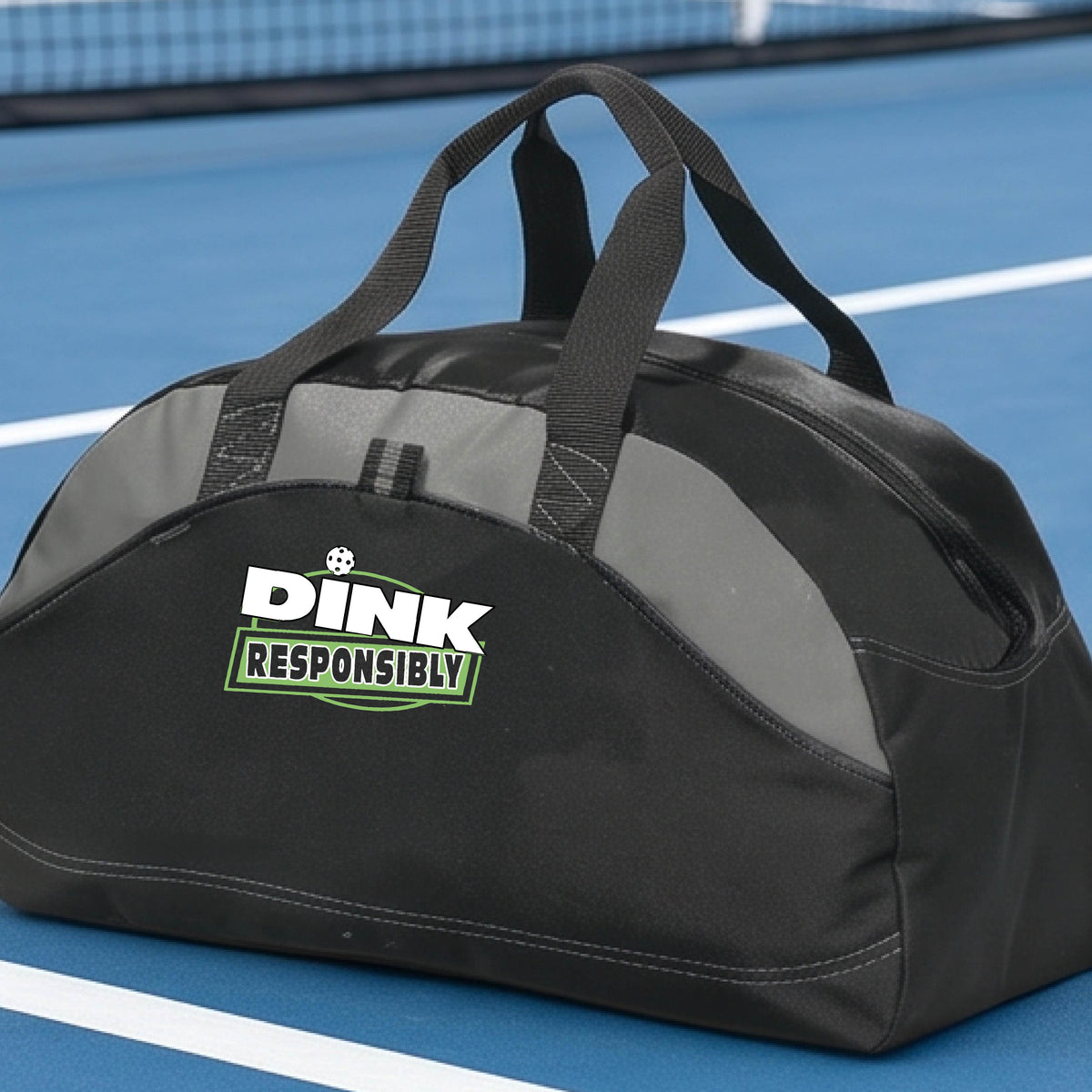 A black, and gray duffel bag with custom 'DINK RESPONSIBLY' branding, placed on a pickleball court with a blue court visible in the background.