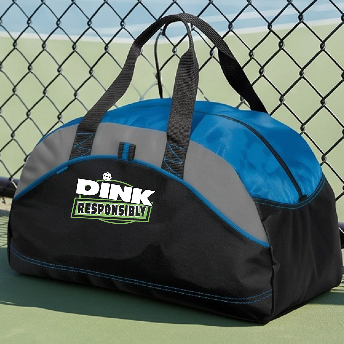A blue, black, and gray duffel bag with custom 'DINK RESPONSIBLY' branding, placed on a pickleball court with a blue court visible in the background.