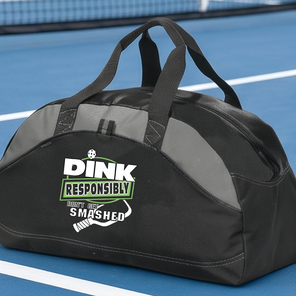 A black, and gray duffel bag with gray panels, featuring the text 'DINK RESPONSIBLY DINK SMASHED' with a white pickleball paddle on the front, on a pickleball court.  