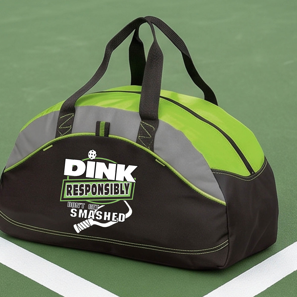A black, gray and lime green duffel bag with gray panels, featuring the text 'DINK RESPONSIBLY DINK SMASHED' with a white pickleball paddle on the front, on a pickleball court.  