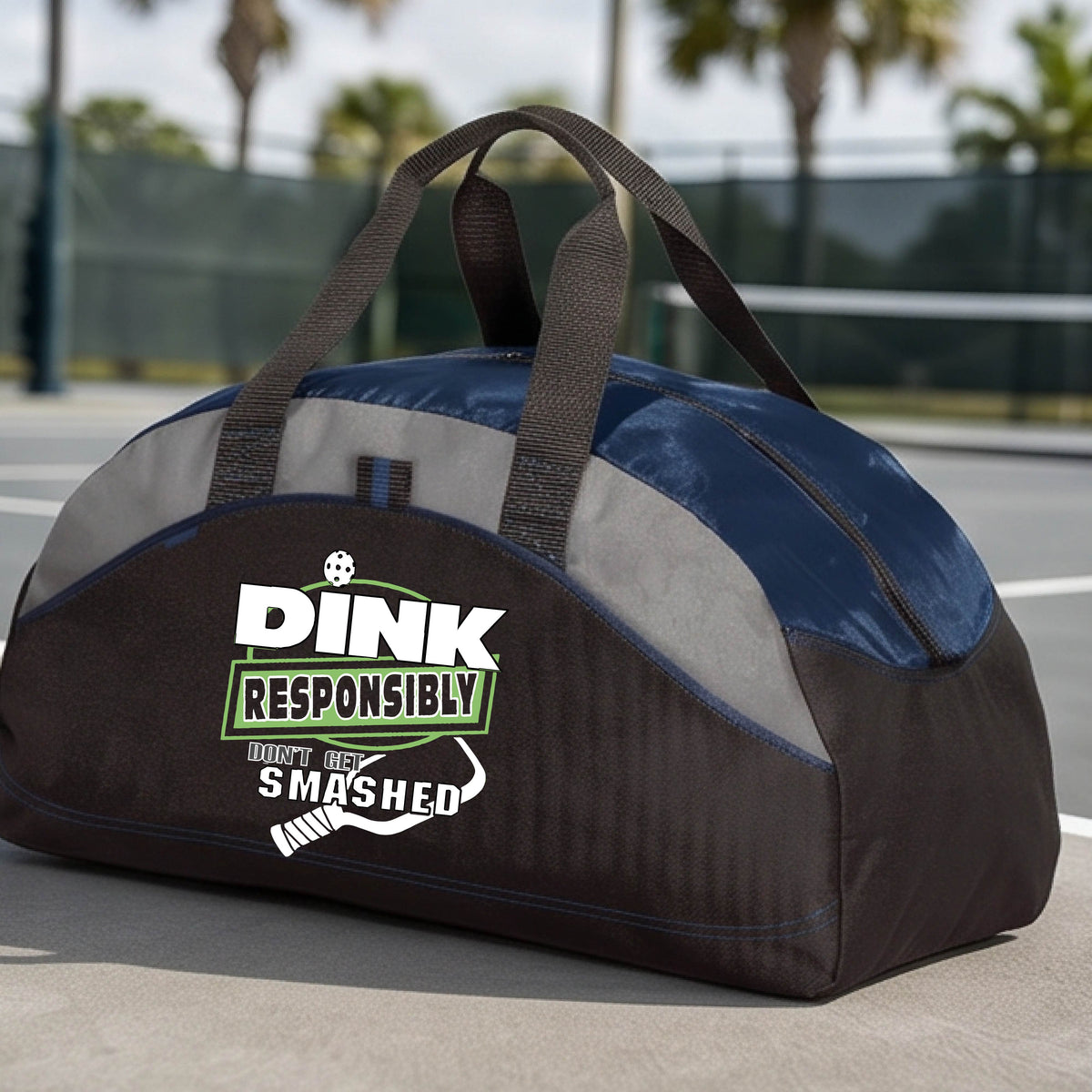 A black, gray and navy blue duffel bag featuring the text 'DINK RESPONSIBLY DINK SMASHED' with a white pickleball paddle on the front, on a pickleball court.  