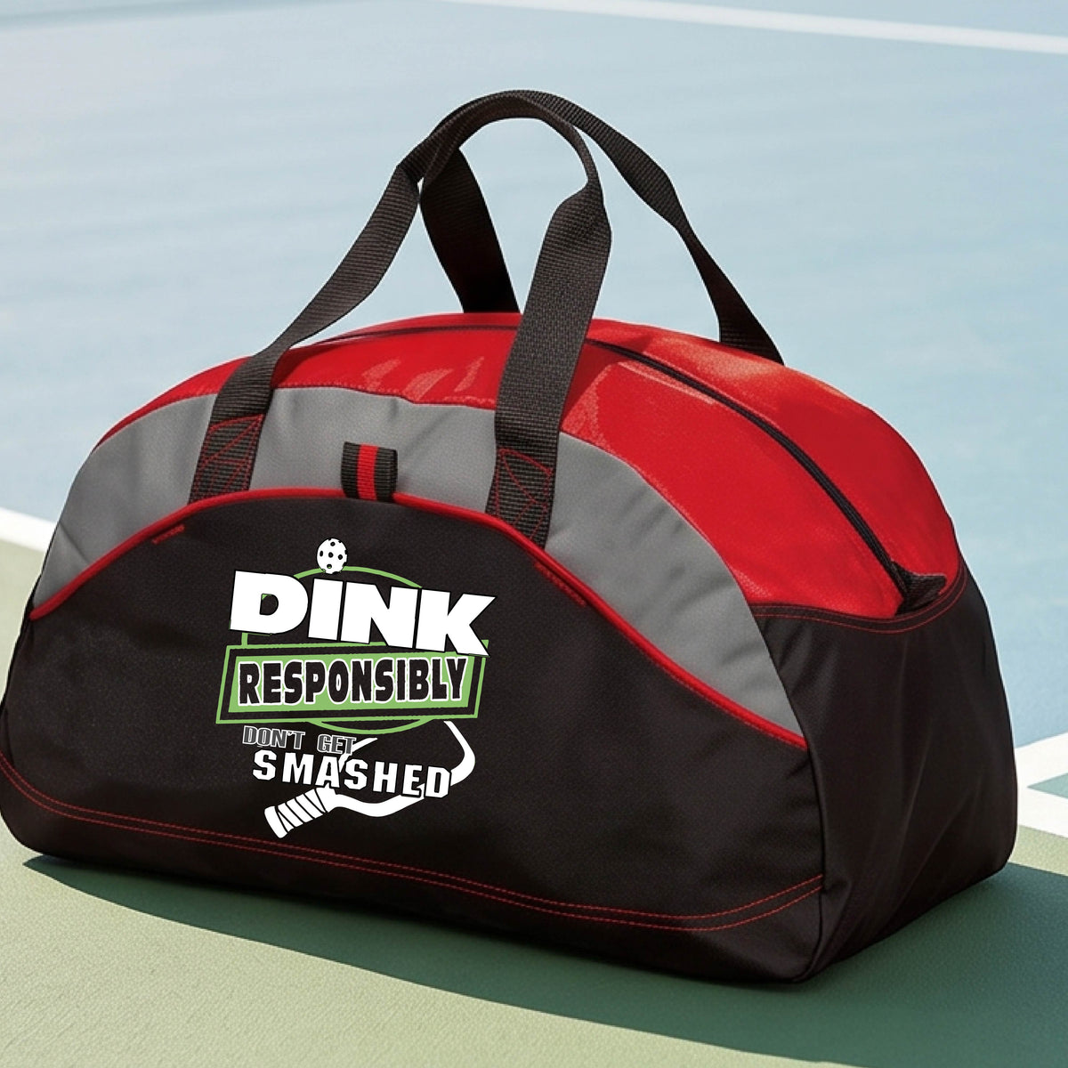A black, gray and red duffel bag featuring the text 'DINK RESPONSIBLY DINK SMASHED' with a white pickleball paddle on the front, on a pickleball court.  