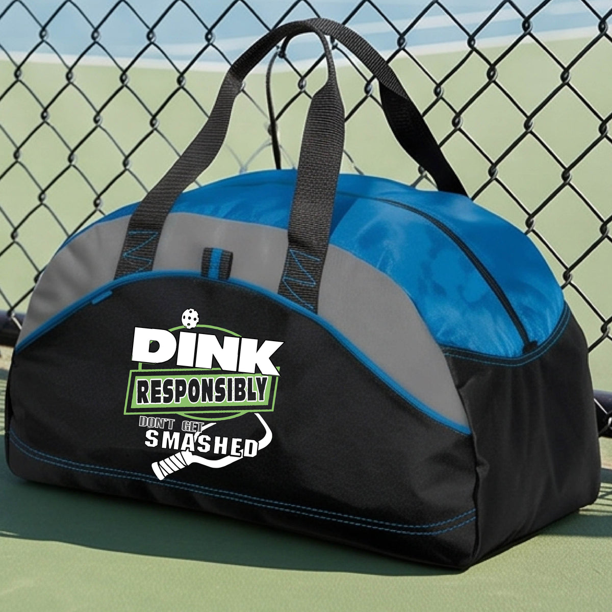 A black, gray and royal blue duffel bag featuring the text 'DINK RESPONSIBLY DINK SMASHED' with a white pickleball paddle on the front, on a pickleball court.  