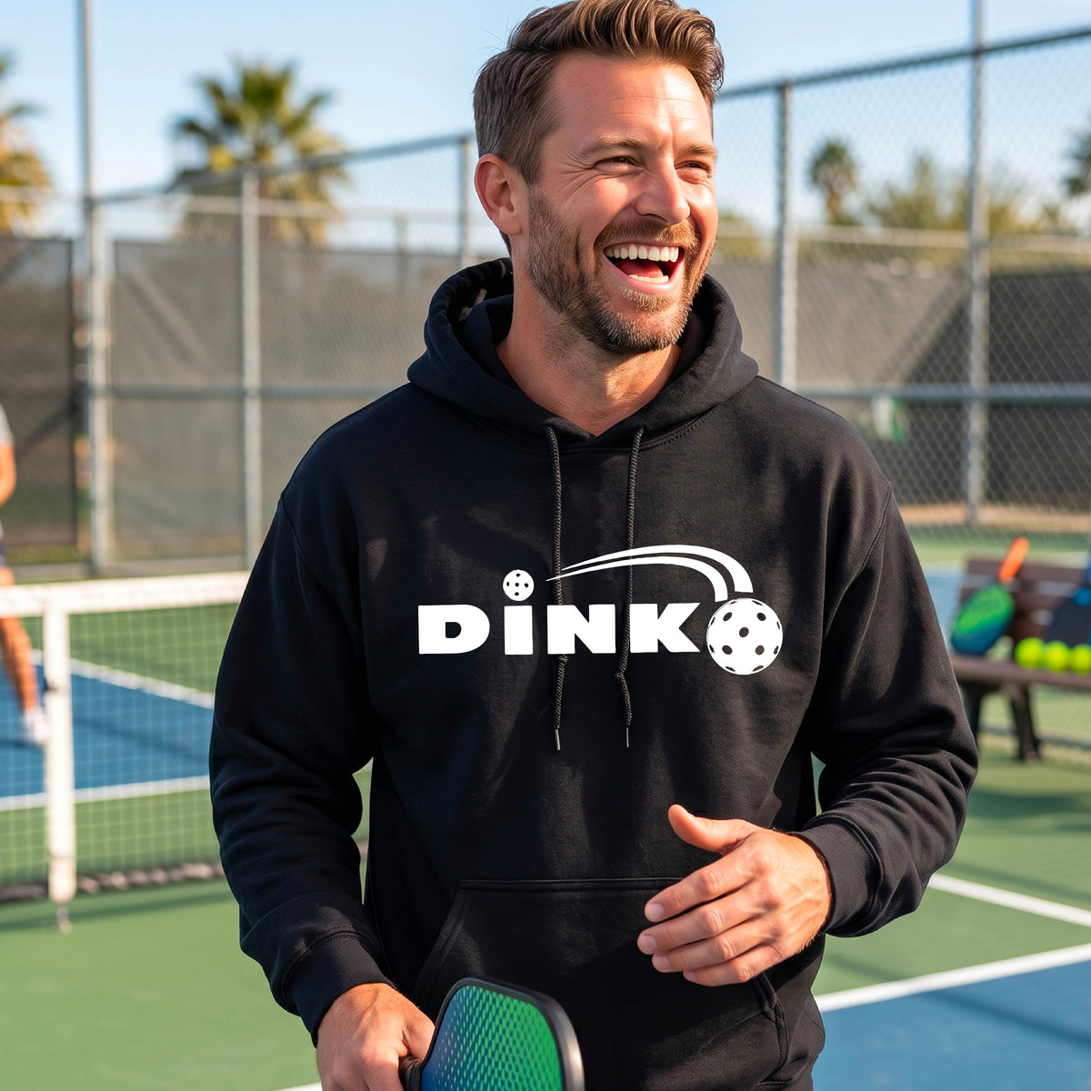 A smiling man wearing a black hoodie with a white 'DINK' graphic featuring a pickleball in flight  set against a blurred pickleball court background.