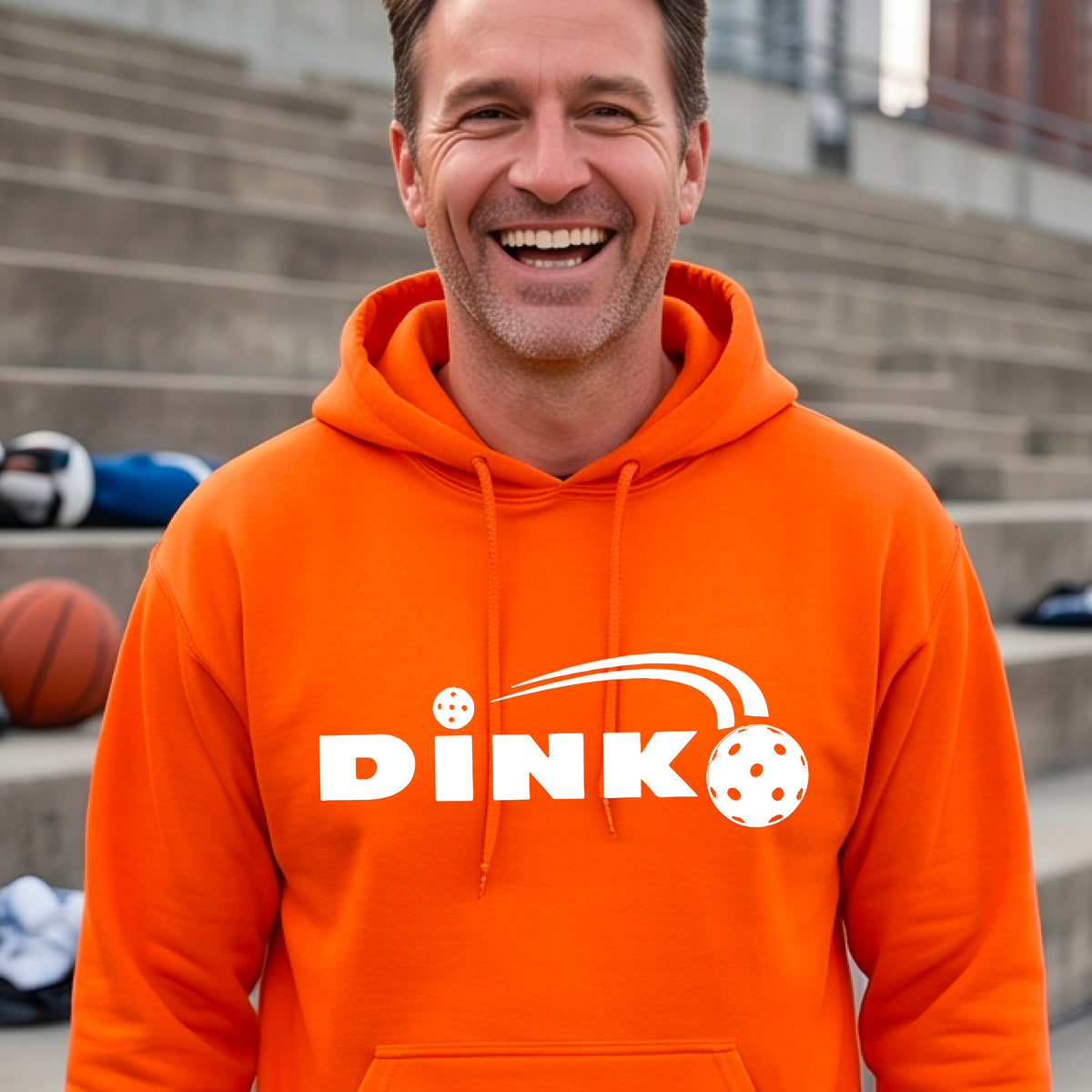 A smiling man wearing an orange hoodie with a white 'DINK' graphic featuring a pickleball in flight  set against a blurred pickleball court background.