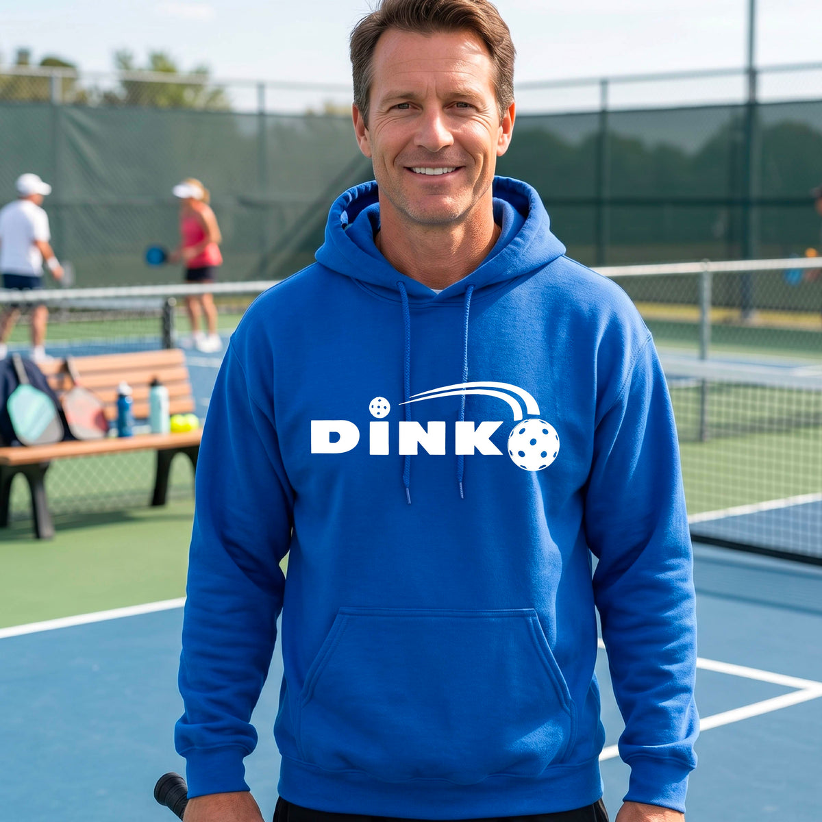 A smiling man wearing a royal blue hoodie with a white 'DINK' graphic featuring a pickleball in flight  set against a blurred pickleball court background.