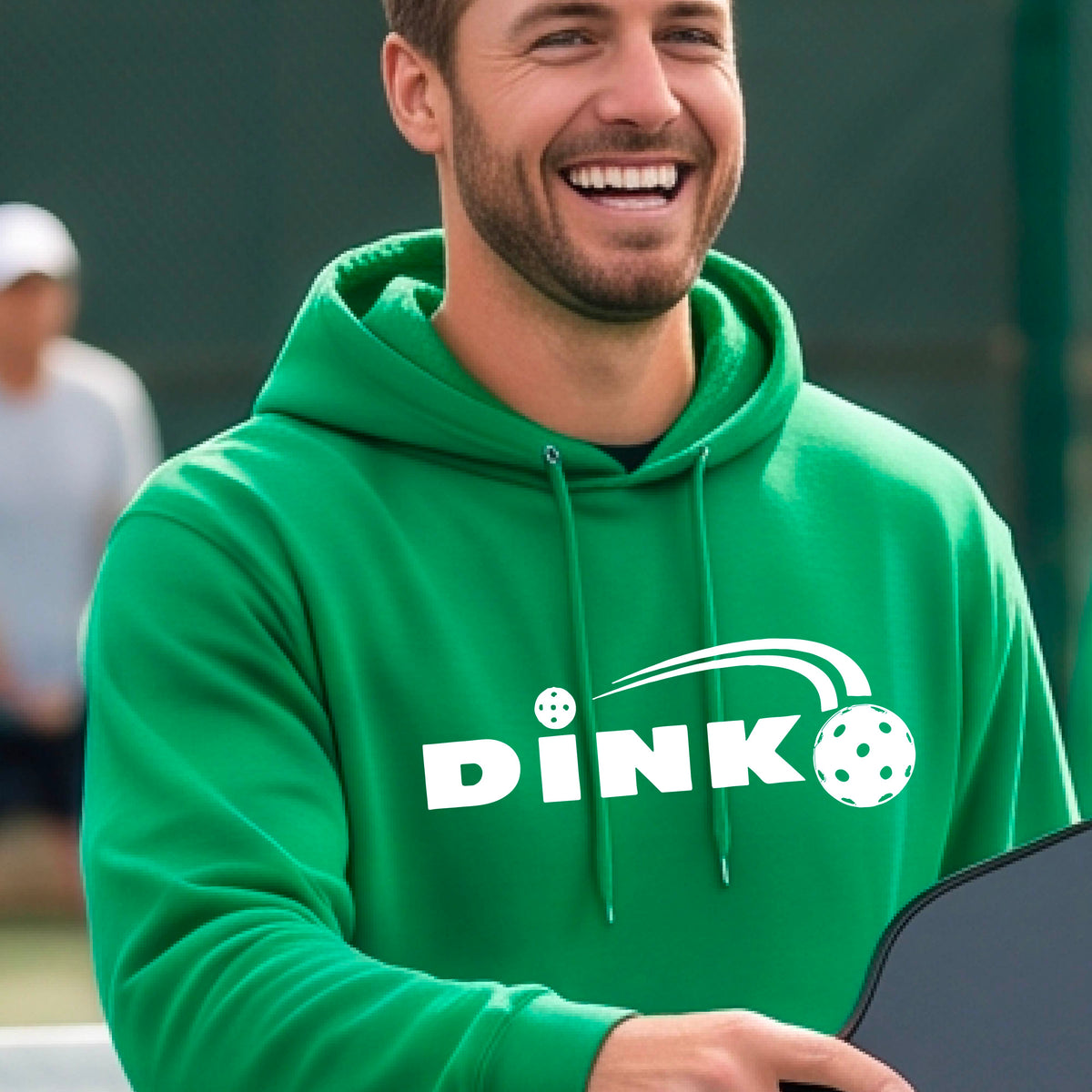 A smiling man wearing a green hoodie with a white 'DINK' graphic featuring a pickleball in flight  set against a blurred pickleball court background.