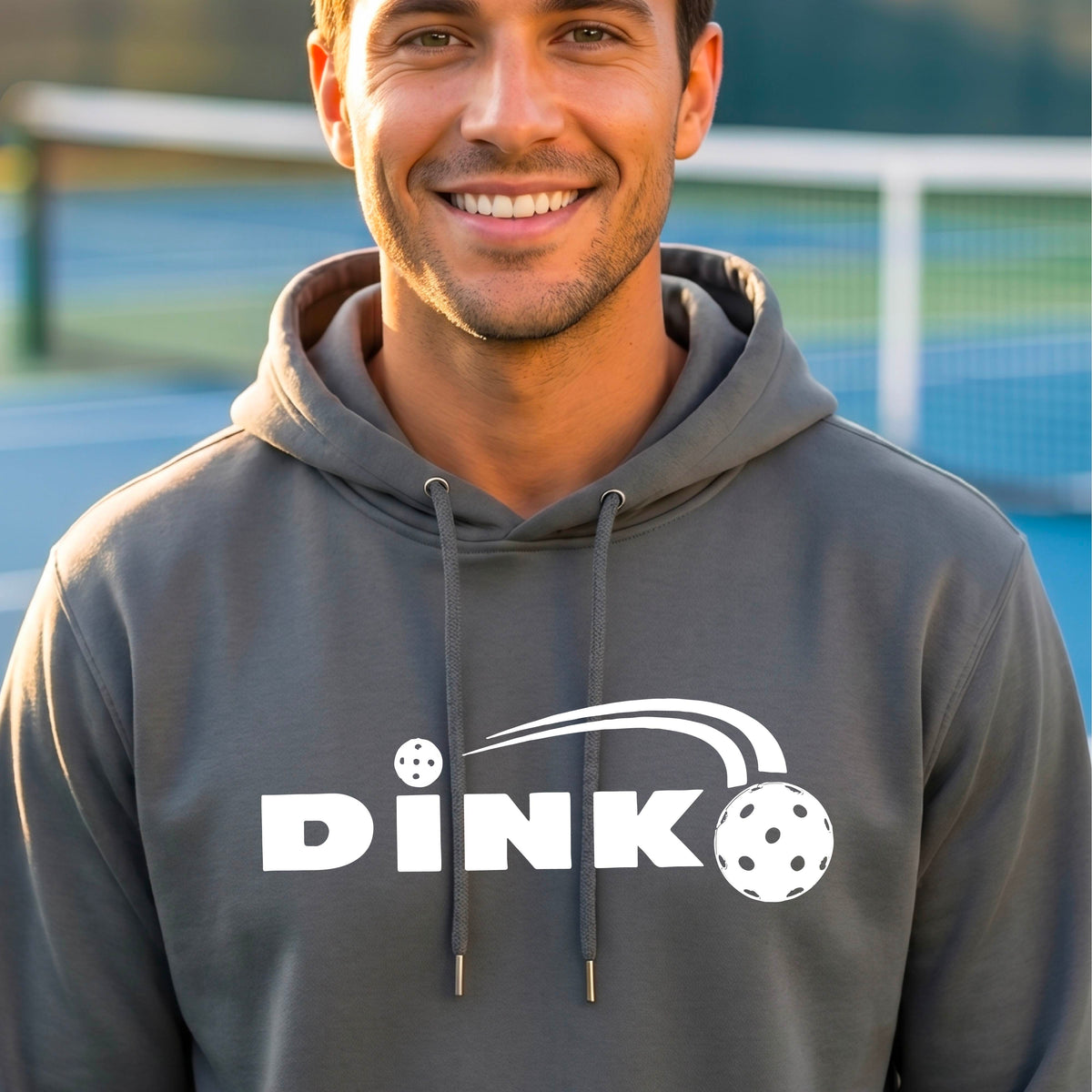 A smiling man wearing a gray hoodie with a white 'DINK' graphic featuring a pickleball in flight  set against a blurred pickleball court background.