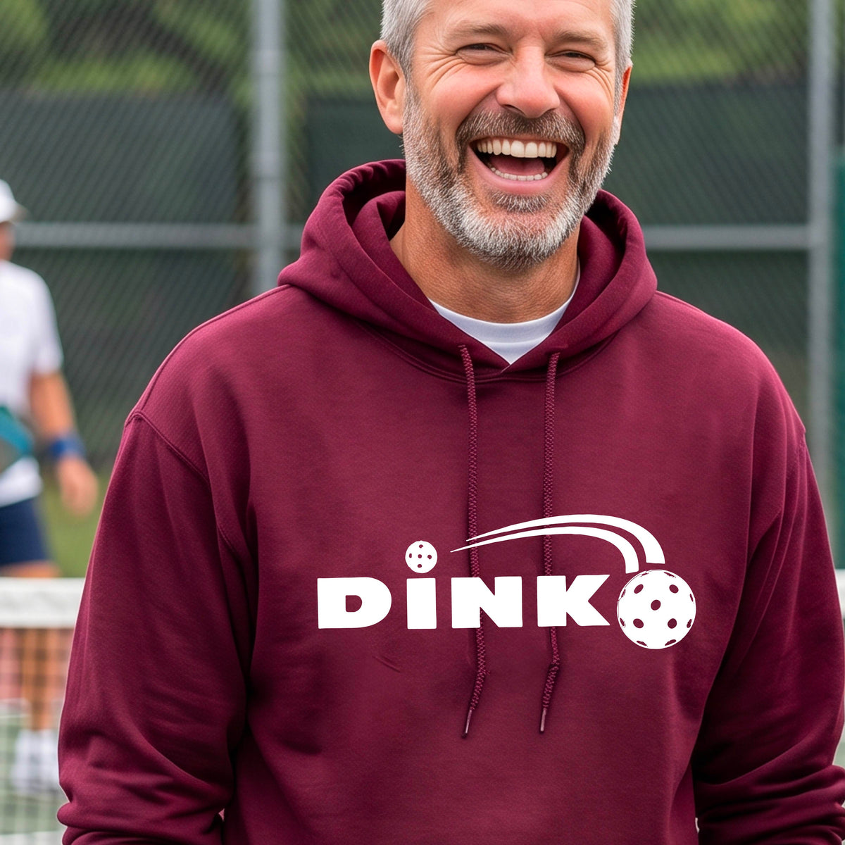 A smiling man wearing a maroon hoodie with a white 'DINK' graphic featuring a pickleball in flight  set against a blurred pickleball court background.