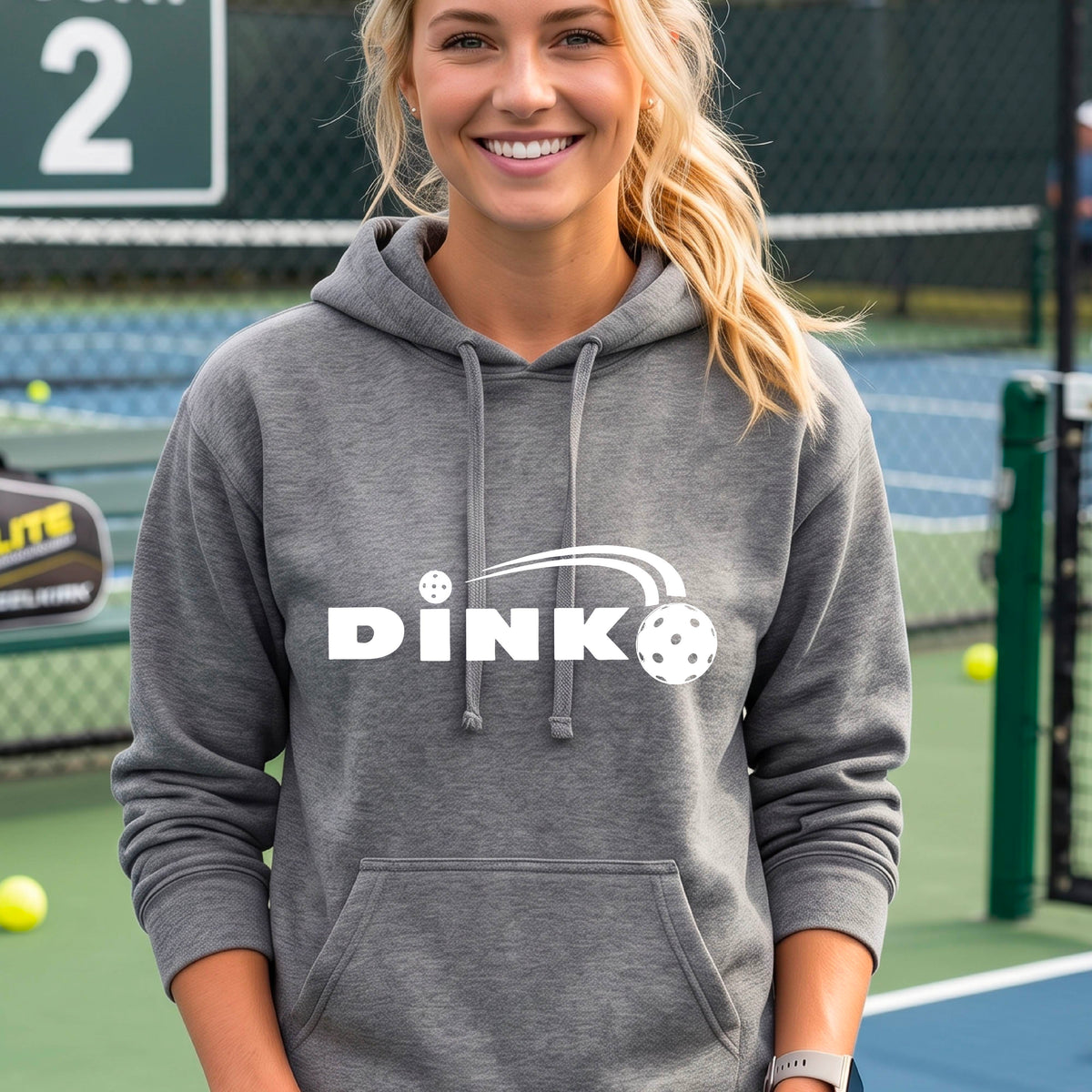 A woman wearing a gray pullover hoodie with a white 'DINK' graphic and a pickleball in flight on the chest, set against a blurred outdoor pickleball background.