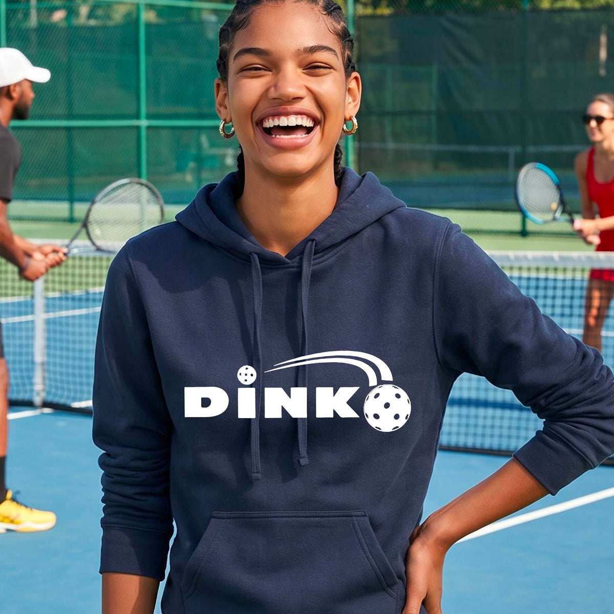 A woman wearing a navy blue pullover hoodie with a white 'DINK' graphic and a pickleball in flight on the chest, set against a blurred outdoor pickleball background.