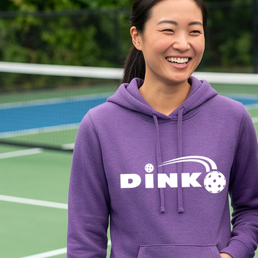 A woman wearing a purple pullover hoodie with a white 'DINK' graphic and a pickleball in flight on the chest, set against a blurred outdoor pickleball background.
