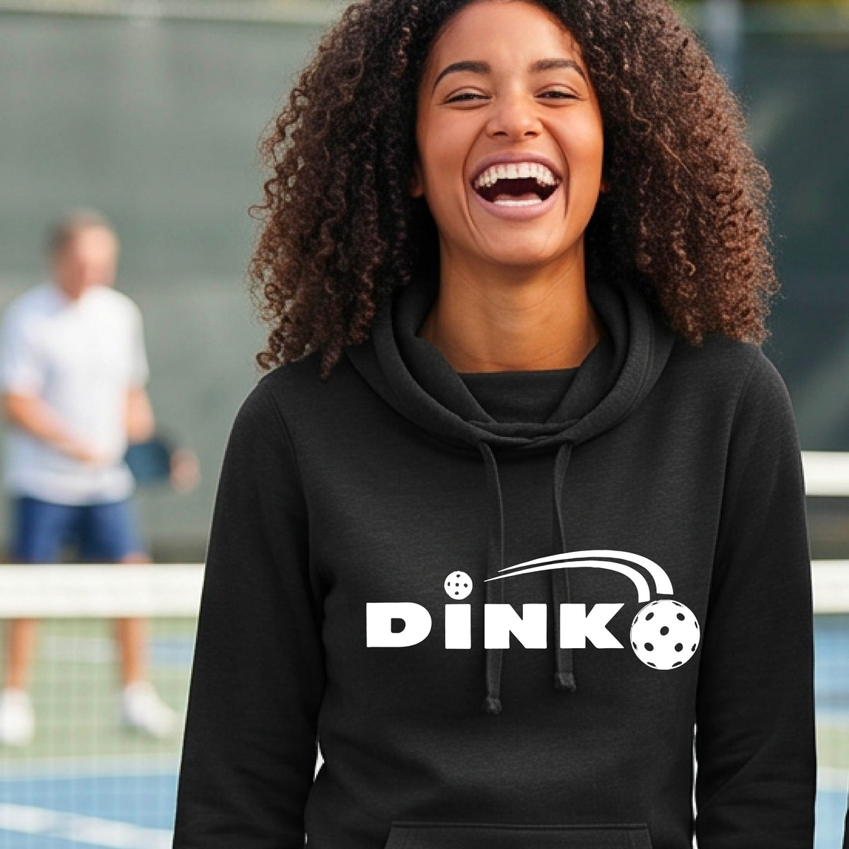 A smiling woman wearing a black cowl-neck pullover hoodie with a white 'DINK' graphic across the chest  and a pickleball in flight with a pickleball court in the background. 