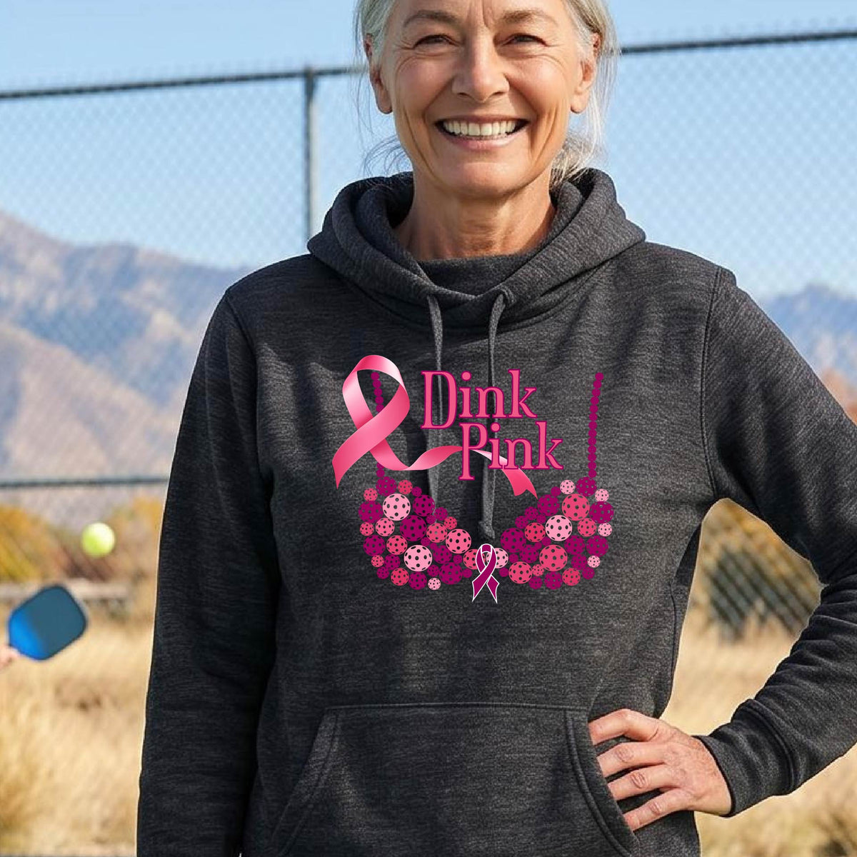 A smiling woman wearing a black cowl-neck hoodie with a pink 'Dink Pink' breast cancer awareness graphic and floral heart design, made of pickleballs and a pink ribbon. 