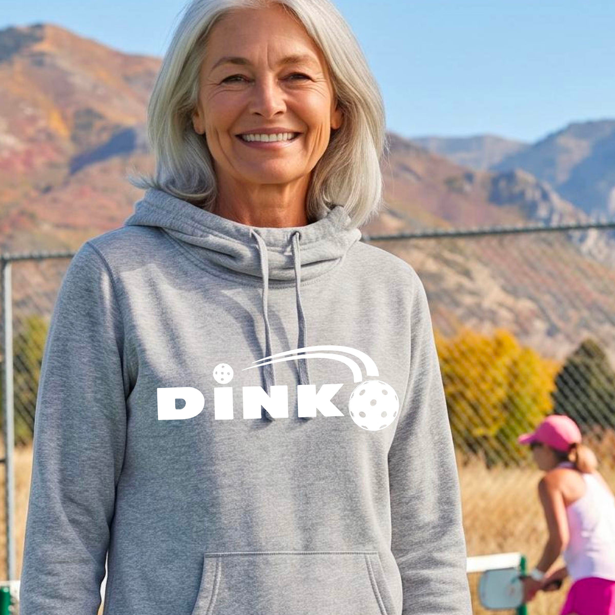 A smiling woman wearing a  gray cowl-neck pullover hoodie with a white 'DINK' graphic across the chest  and a pickleball in flight with a pickleball court in the background. 