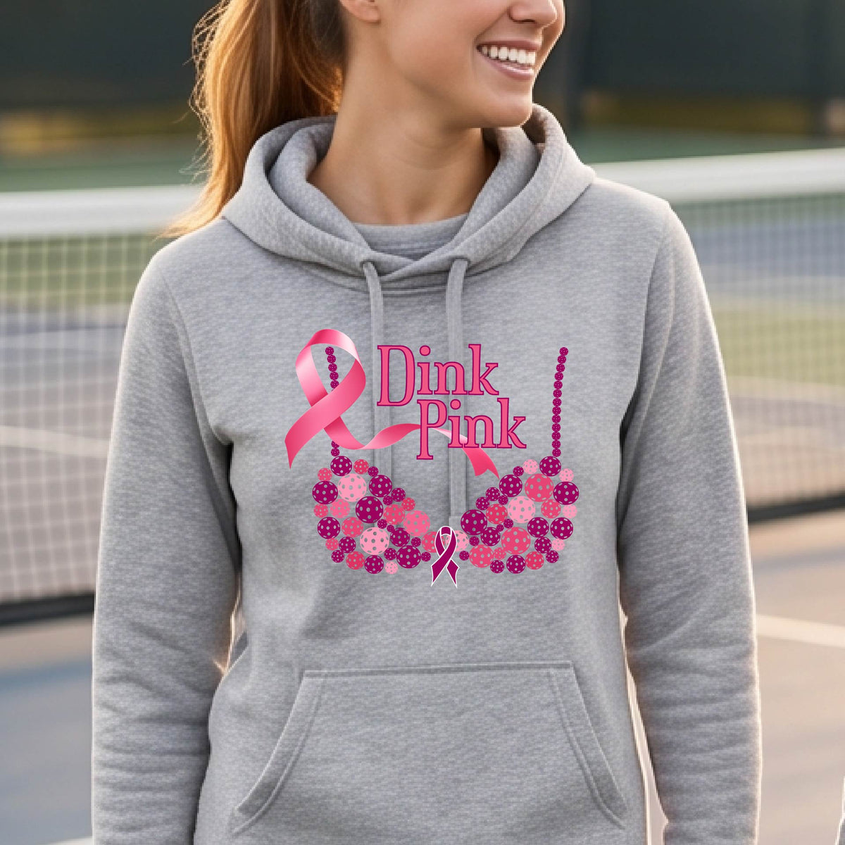 A smiling woman wearing a light gray cowl-neck hoodie with a pink 'Dink Pink' breast cancer awareness graphic and floral heart design, made of pickleballs and a pink ribbon. 