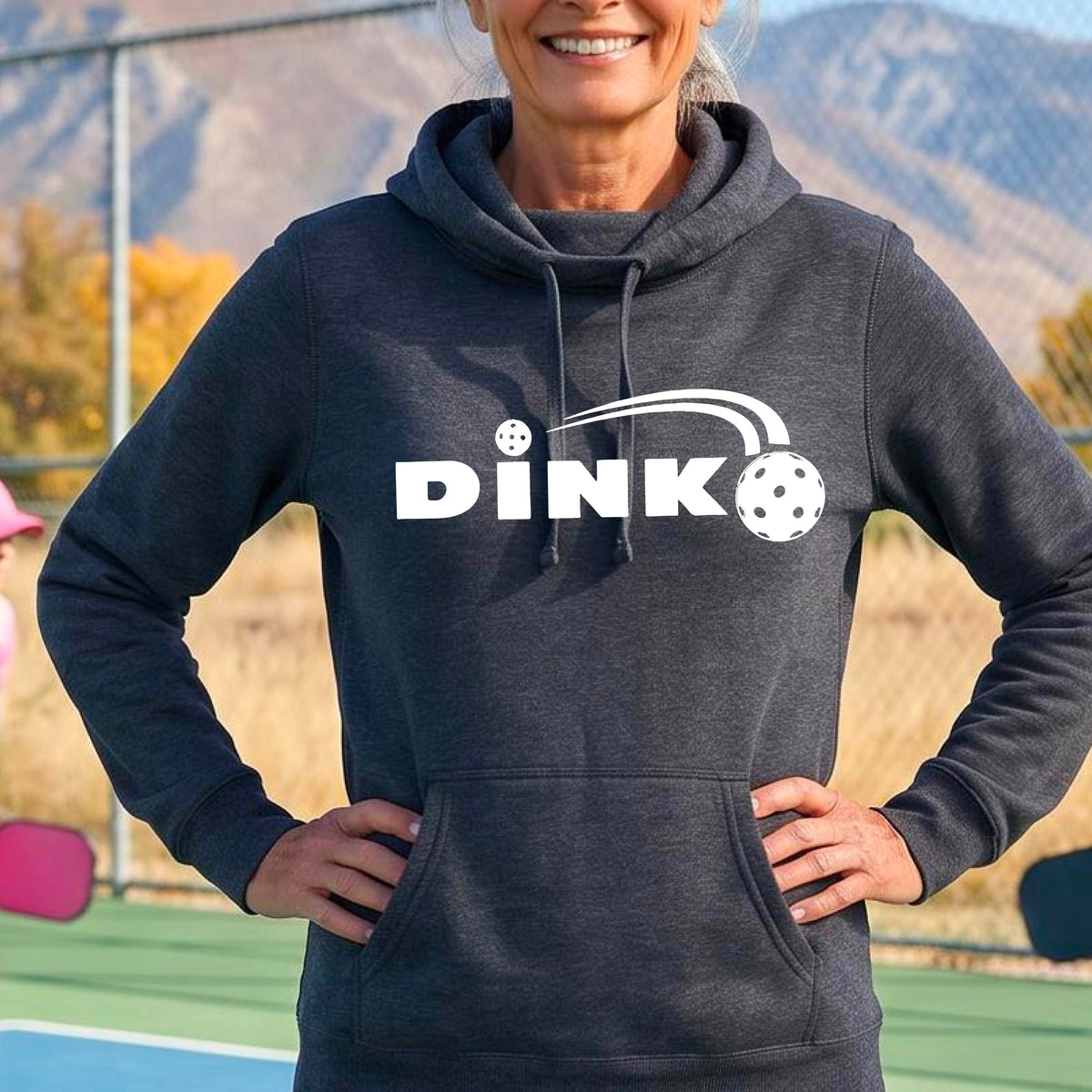 A smiling woman wearing a navy blue cowl-neck pullover hoodie with a white 'DINK' graphic across the chest  and a pickleball in flight with a pickleball court in the background. 