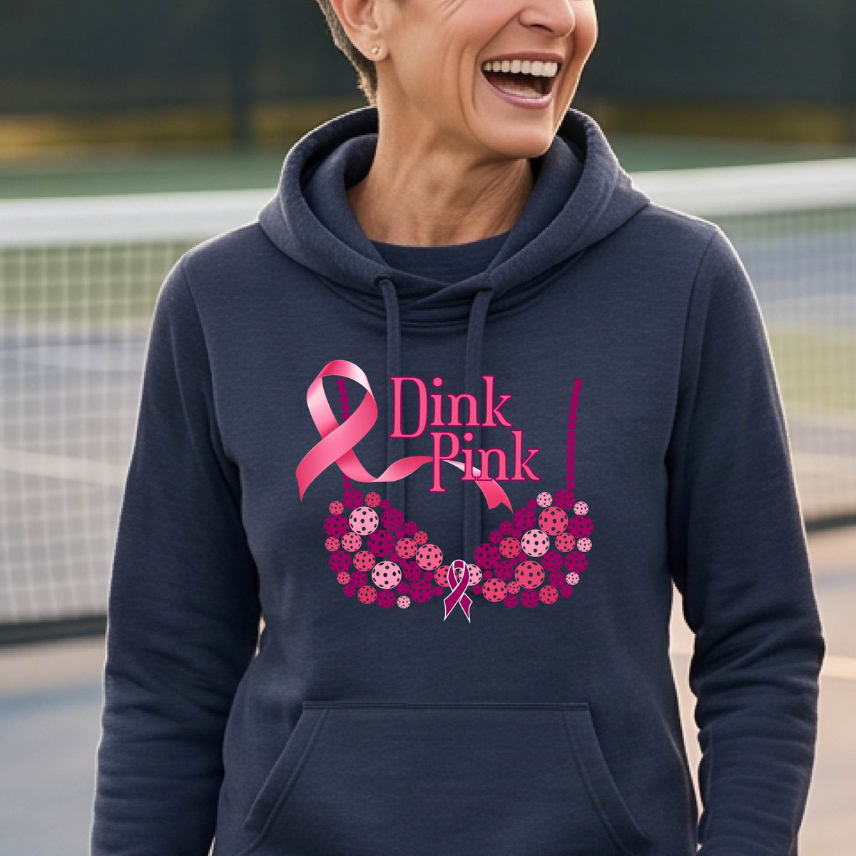 A smiling woman wearing a navy blue cowl-neck hoodie with a pink 'Dink Pink' breast cancer awareness graphic and floral heart design, made of pickleballs and a pink ribbon. 