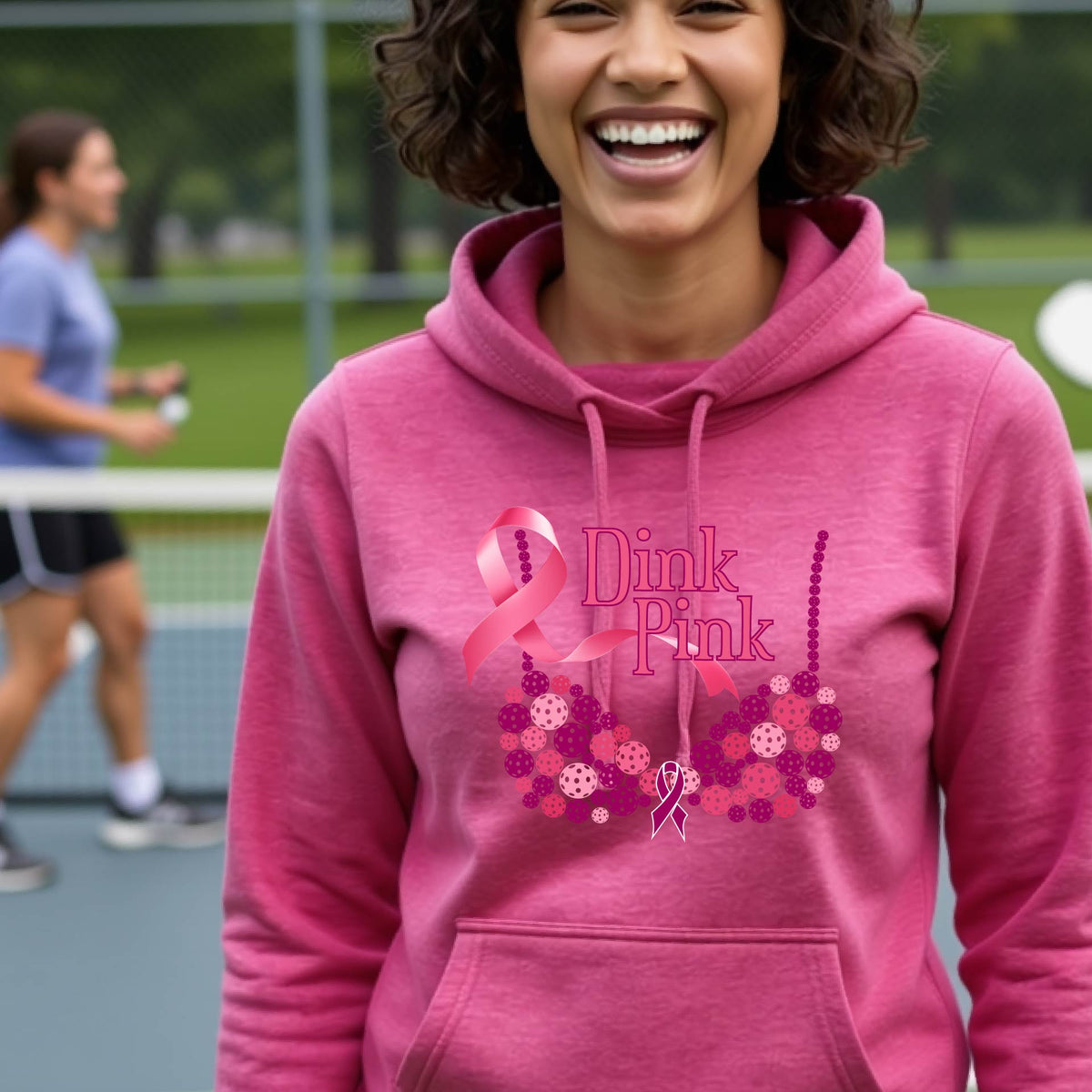 A smiling woman wearing a pink cowl-neck hoodie with a pink 'Dink Pink' breast cancer awareness graphic and floral heart design, made of pickleballs and a pink ribbon. 