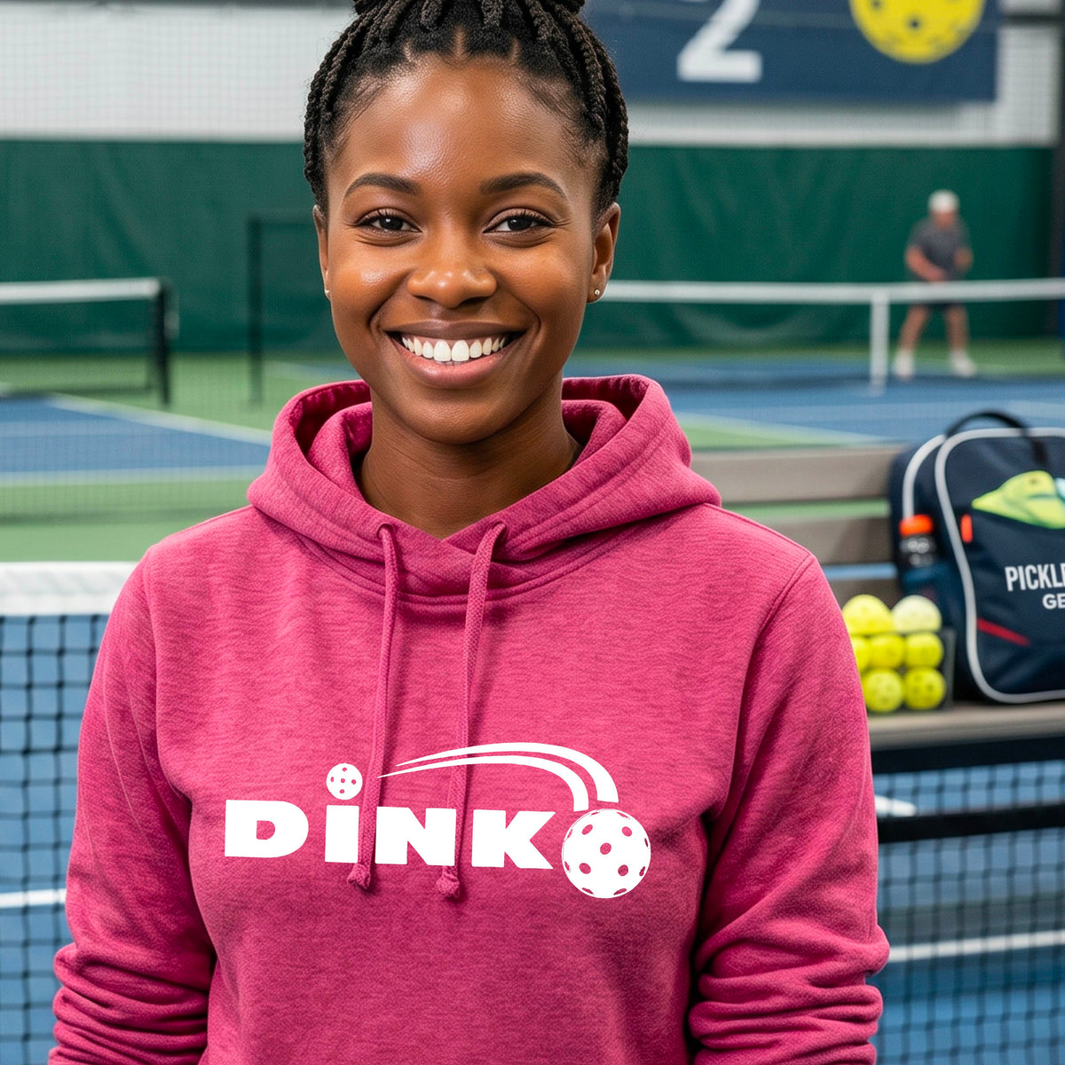 A smiling woman wearing a pink cowl-neck pullover hoodie with a white 'DINK' graphic across the chest  and a pickleball in flight with a pickleball court in the background. 