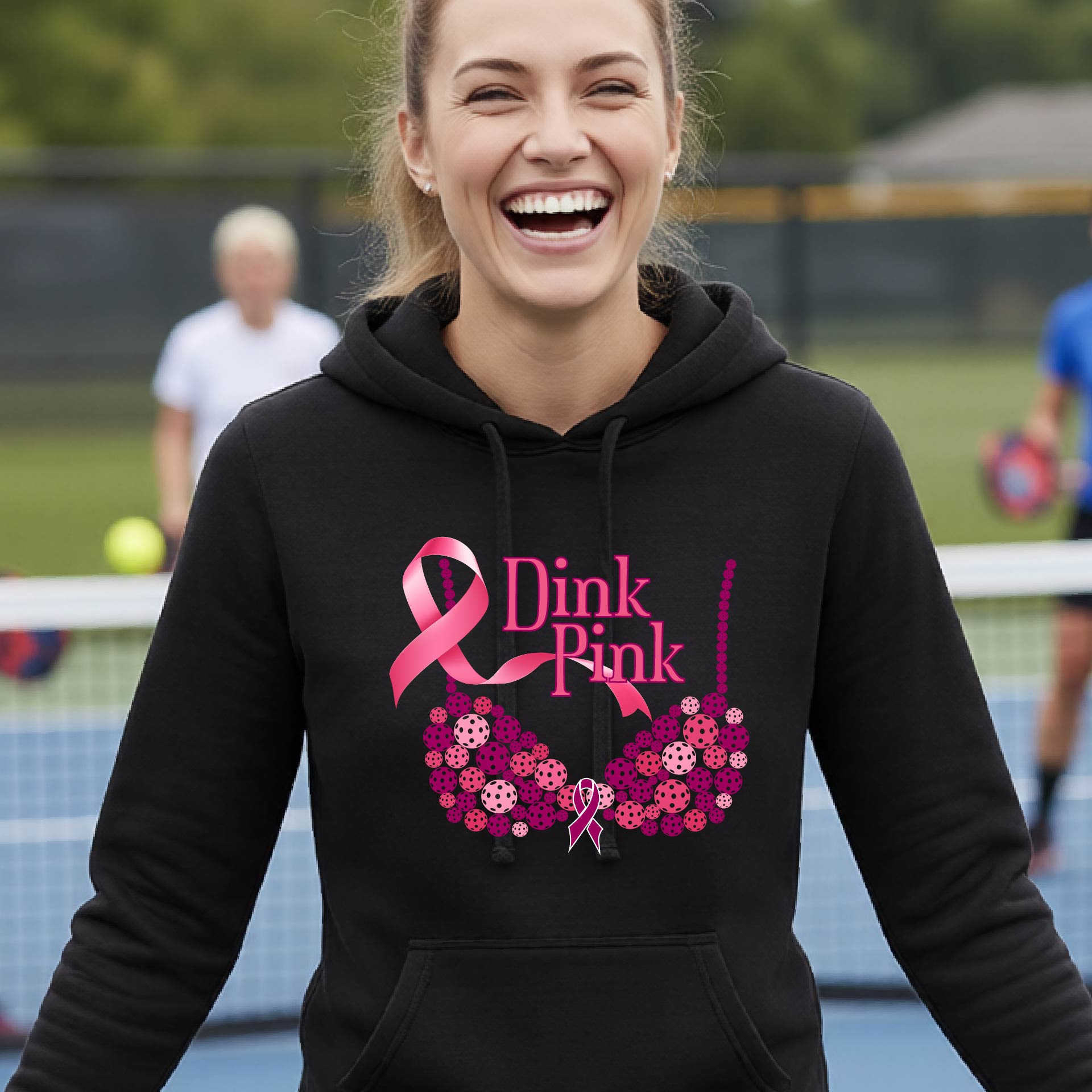 A woman smiling on a pickleball court wearing a black hoodie with a pink 'Dink Pink' graphic made of pickleball and a ribbon and a bra-shaped floral design.